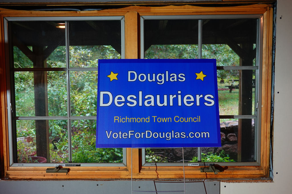 A blue yard sign with my name on it in front of window with greenery behind it